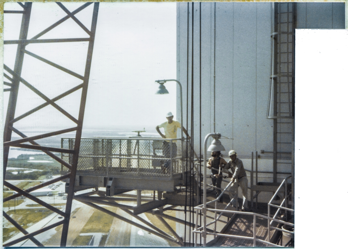 Image 098. Viewed looking south from the Fixed Service Structure at Space Shuttle Launch Complex 39-B, Kennedy Space Center, Florida, left-to-right, James MacLaren, Tommy Northcutt, and Carlton D. Taylor can be seen taking a lighter moment, posing for a picture at their place of work, on the Left SRB Access Platform and Catwalk of the Rotating Service Structure, with the RCS Room behind them to their right, and the foot of the Pad Slope, the OPS Building, the Crawlerway, and the wilderness of the Merritt Island Wildlife Refuge in the distance to their left. MacLaren wrote and photographed these essays, but on this occasion handed his camera off to Jack Petty, ex-ironworker who worked for BRPH as Structural Tech Rep during the 79K24048 portions of work on Pad B. Tommy Northcutt was a Union Ironworker who worked for Ivey Steel at the same time, and was a Good Man, utterly without guile or pretense, kind-natured, and an excellent ironworker. Carlton D. Taylor worked Contract Oversight as a Field Representative and was also a Good Man, with a wicked sense of humor, but who also tended toward hyperbole, especially when speaking of his own exploits, and Jack Petty hung the moniker of ”Ten Percent Taylor,” on him, only half in jest, which he would invariably follow up with, ”You can believe ten percent of everything he tells you,” followed by a good-natured laugh. The three of us, MacLaren, Petty, and Taylor, bonded closely for the duration of the project, and formed an excellent working team, one from the Contractor (MacLaren), one from the A&E (Petty), and one from Contracts (Taylor). We trusted each other completely, and cut through a tremendous amount of bullshit, getting a very large amount of work done by dispensing with anything at all which smacked of delaying formality or mistrust. It was a Good Team.  Photo by Jack Petty.